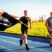 Two men in athletic wear are running on a blue track, with one carrying a black POW/MIA flag on a wooden pole. The flag is billowing in the wind, displaying the recognizable POW/MIA silhouette and the text 'YOU ARE NOT FORGOTTEN' (visible in reverse due to wind direction). The setting appears to be an outdoor athletic facility during golden hour lighting conditions.