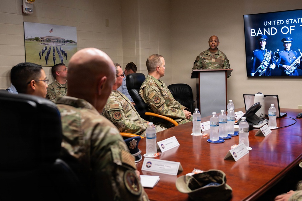 A military briefing or presentation is taking place in a conference room, with a uniformed officer speaking at a podium to a group of seated military personnel. A television screen behind the speaker displays a partially visible slide reading 'United States Band of T[he...]', suggesting a presentation about the United States Air Force Band. Name placards are visible on the conference table along with water bottles.