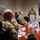 A military briefing or presentation is taking place in a conference room, with a uniformed officer speaking at a podium to a group of seated military personnel. A television screen behind the speaker displays a partially visible slide reading 'United States Band of T[he...]', suggesting a presentation about the United States Air Force Band. Name placards are visible on the conference table along with water bottles.