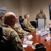 A military briefing or presentation is taking place in a conference room, with a uniformed officer speaking at a podium to a group of seated military personnel. A television screen behind the speaker displays a partially visible slide reading 'United States Band of T[he...]', suggesting a presentation about the United States Air Force Band. Name placards are visible on the conference table along with water bottles.