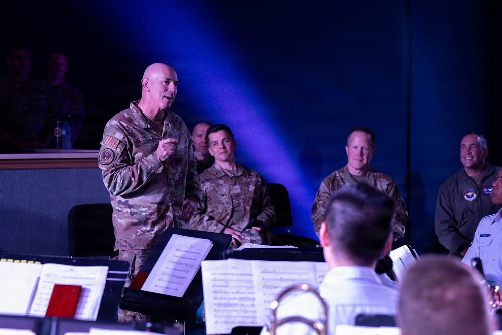 A senior military officer in OCP (Operational Camouflage Pattern) uniform stands at the front of what appears to be a military band performance, addressing musicians with sheet music visible on stands. Several other military personnel in various uniforms are seated in the background, appearing attentive and engaged. The setting appears to be an indoor performance venue with dramatic blue stage lighting.