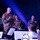 A senior military officer in OCP (Operational Camouflage Pattern) uniform stands at the front of what appears to be a military band performance, addressing musicians with sheet music visible on stands. Several other military personnel in various uniforms are seated in the background, appearing attentive and engaged. The setting appears to be an indoor performance venue with dramatic blue stage lighting.