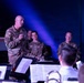 A senior military officer in OCP (Operational Camouflage Pattern) uniform stands at the front of what appears to be a military band performance, addressing musicians with sheet music visible on stands. Several other military personnel in various uniforms are seated in the background, appearing attentive and engaged. The setting appears to be an indoor performance venue with dramatic blue stage lighting.