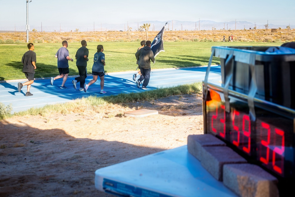 A group of approximately five runners are jogging on a blue outdoor track in what appears to be a desert or arid environment. One runner is carrying a dark flag or banner. A digital timer in the foreground displays the time 23:59:32, suggesting this is a timed running event or physical fitness test.