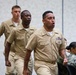 Three U.S. Navy sailors in tan/khaki service uniforms are walking in formation, appearing to be reciting or chanting something aloud. The lead sailor displays multiple ribbons, a warfare device, and a submarine or surface warfare badge on his chest. In the background, two additional service members in Navy Working Uniform (NWU) camouflage are seated.
