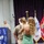 A military promotion or pinning ceremony is taking place in a formal venue. A woman in a green dress is holding a young child who appears to be placing or touching a cover (military hat) on a uniformed Navy service member's head. A second uniformed individual stands at a podium in the background, smiling.