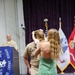 A military promotion or pinning ceremony is taking place in a formal venue. A woman in a green dress is holding a young child who appears to be placing or touching a cover (military hat) on a uniformed Navy service member's head. A second uniformed individual stands at a podium in the background, smiling.