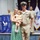 A military ceremony featuring a U.S. Navy Chief Petty Officer in Service Khaki uniform posing with a woman in a green floral dress holding a young toddler girl. The setting appears to be an official CPO (Chief Petty Officer) selectee ceremony, as indicated by the blue banner reading 'Space Coast CPO FY27 Selectees' in the background. Additional Navy personnel and an Army flag are visible in the background.