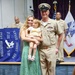 A military ceremony featuring a U.S. Navy Chief Petty Officer in Service Khaki uniform posing with a woman in a green floral dress holding a young toddler girl. The setting appears to be an official CPO (Chief Petty Officer) selectee ceremony, as indicated by the blue banner reading 'Space Coast CPO FY27 Selectees' in the background. Additional Navy personnel and an Army flag are visible in the background.