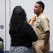 A military promotion or award ceremony is taking place, with a woman in civilian clothing pinning something onto the collar or shoulder area of a U.S. Navy sailor in khaki service uniform. The sailor displays multiple ribbons, a warfare device, and a command insignia on his uniform. Seated observers in Navy working uniforms are visible in the background.