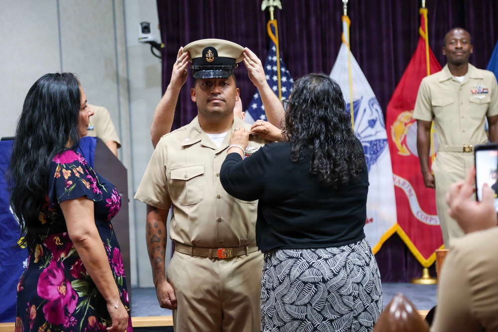 A military promotion ceremony is taking place, likely a U.S. Navy Chief Petty Officer pinning/hat ceremony. Two women are placing a chief petty officer's combination cover (hat) on a male sailor in khaki uniform, while another uniformed sailor stands in the background. The event appears to be held in a formal indoor venue decorated with military flags including the U.S. flag and Marine Corps flag.