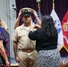 A military promotion ceremony is taking place, likely a U.S. Navy Chief Petty Officer pinning/hat ceremony. Two women are placing a chief petty officer's combination cover (hat) on a male sailor in khaki uniform, while another uniformed sailor stands in the background. The event appears to be held in a formal indoor venue decorated with military flags including the U.S. flag and Marine Corps flag.