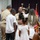 A military promotion or pinning ceremony is taking place in a formal indoor setting. A service member in khaki/tan uniform is surrounded by family members, including children dressed in white, who appear to be participating in the pinning of insignia. Military flags and a podium are visible in the background.