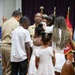A military promotion or pinning ceremony is taking place in a formal indoor setting. A service member in khaki/tan uniform is surrounded by family members, including children dressed in white, who appear to be participating in the pinning of insignia. Military flags and a podium are visible in the background.