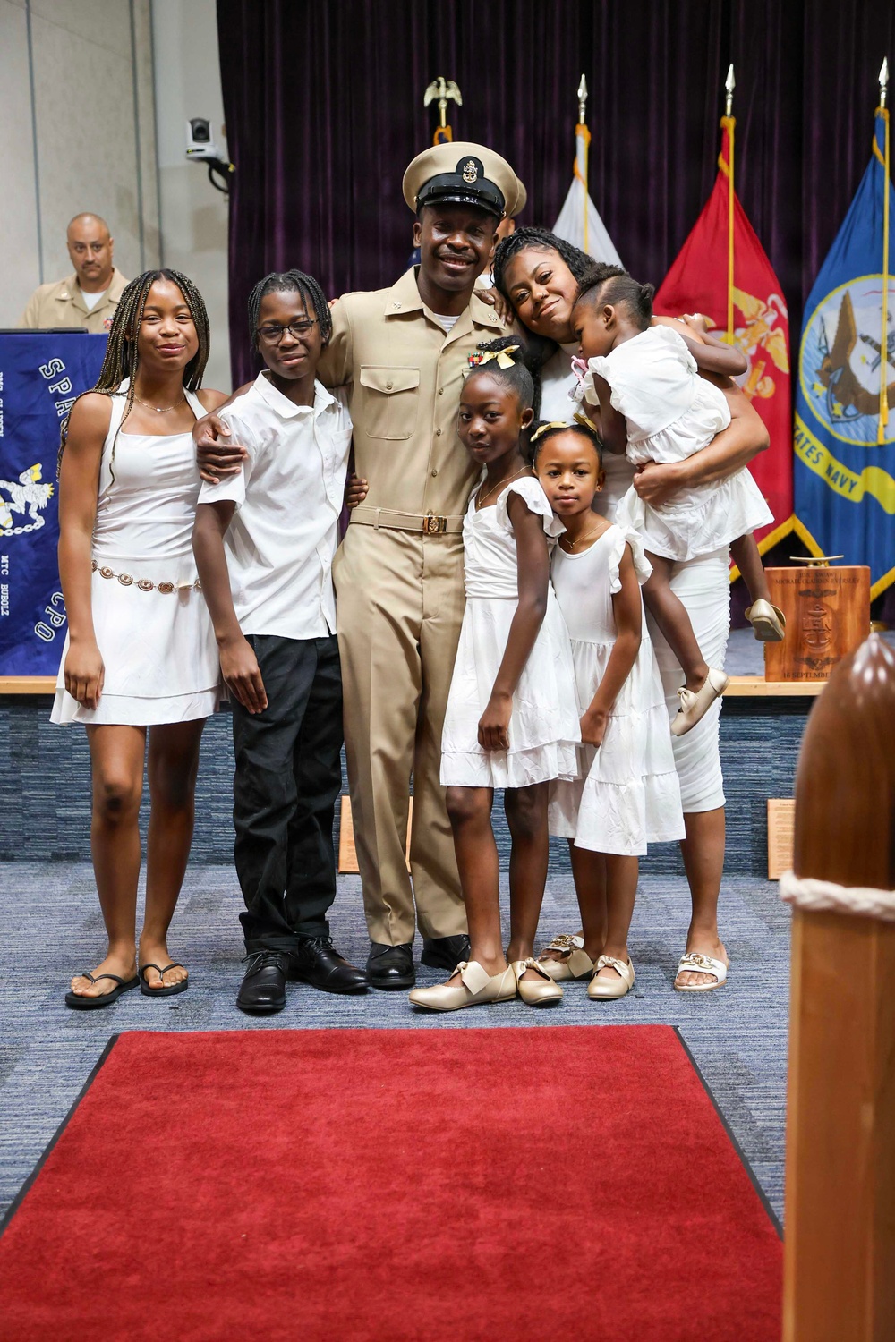 A U.S. Navy Chief Petty Officer in dress khaki uniform poses with his family during what appears to be a promotion or advancement ceremony. The family members, dressed predominantly in white, gather around him in a formal military venue decorated with service flags and a red carpet. The occasion appears celebratory, likely marking a significant military milestone.