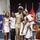 A U.S. Navy Chief Petty Officer in dress khaki uniform poses with his family during what appears to be a promotion or advancement ceremony. The family members, dressed predominantly in white, gather around him in a formal military venue decorated with service flags and a red carpet. The occasion appears celebratory, likely marking a significant military milestone.