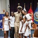 A U.S. Navy Chief Petty Officer in dress khaki uniform poses with his family during what appears to be a promotion or advancement ceremony. The family members, dressed predominantly in white, gather around him in a formal military venue decorated with service flags and a red carpet. The occasion appears celebratory, likely marking a significant military milestone.