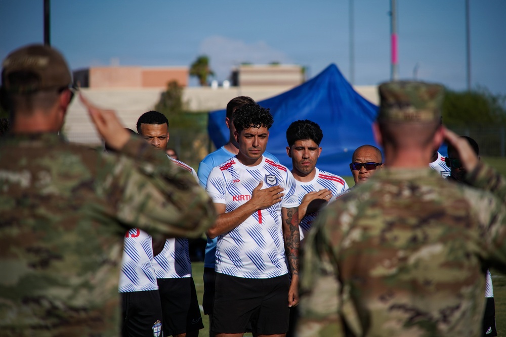 A group of soccer players in white jerseys with blue diagonal stripes stand with their hands over their hearts, appearing to observe a national anthem or ceremonial moment. Two military personnel in camouflage uniforms are visible in the foreground with their backs to the camera, one appearing to salute. The setting appears to be an outdoor sports field with a blue tent and buildings visible in the background.