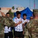 A group of soccer players in white jerseys with blue diagonal stripes stand with their hands over their hearts, appearing to observe a national anthem or ceremonial moment. Two military personnel in camouflage uniforms are visible in the foreground with their backs to the camera, one appearing to salute. The setting appears to be an outdoor sports field with a blue tent and buildings visible in the background.