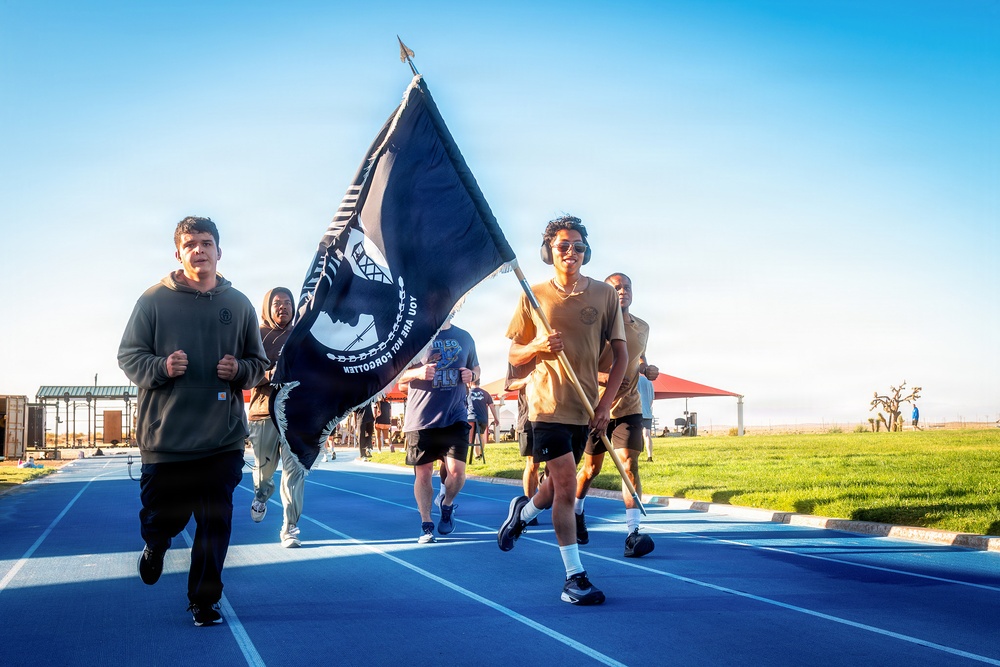 A group of people are running on a blue athletic track while one participant carries a POW/MIA flag. The event appears to be a commemorative run or memorial event held outdoors during daylight hours. The setting includes a well-maintained sports facility with green grass, a pavilion, and clear blue skies.