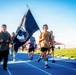 A group of people are running on a blue athletic track while one participant carries a POW/MIA flag. The event appears to be a commemorative run or memorial event held outdoors during daylight hours. The setting includes a well-maintained sports facility with green grass, a pavilion, and clear blue skies.