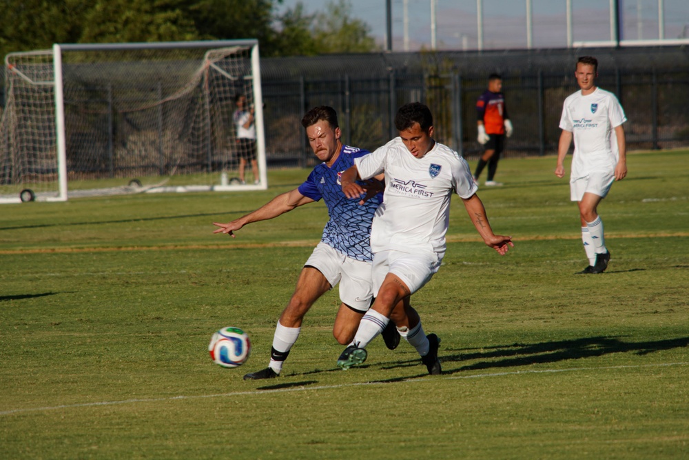 A soccer match is in progress on a grass field, with two players in the foreground competing for the ball in a physical challenge. One player wears a blue patterned jersey while the other wears a white jersey with 'America First' sponsorship. Additional players, a goalkeeper, and a goal post are visible in the background.