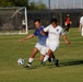A soccer match is in progress on a grass field, with two players in the foreground competing for the ball in a physical challenge. One player wears a blue patterned jersey while the other wears a white jersey with 'America First' sponsorship. Additional players, a goalkeeper, and a goal post are visible in the background.