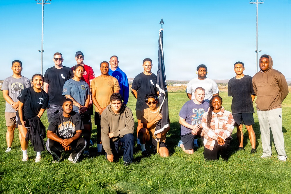 A group of approximately 16 people pose together on a grass field for a group photo. One person in the front row is holding a dark pennant or guidon flag with a spear-tip finial. The setting appears to be an outdoor athletic or recreational field with stadium light poles visible in the background under a clear blue sky.