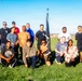 A group of approximately 16 people pose together on a grass field for a group photo. One person in the front row is holding a dark pennant or guidon flag with a spear-tip finial. The setting appears to be an outdoor athletic or recreational field with stadium light poles visible in the background under a clear blue sky.