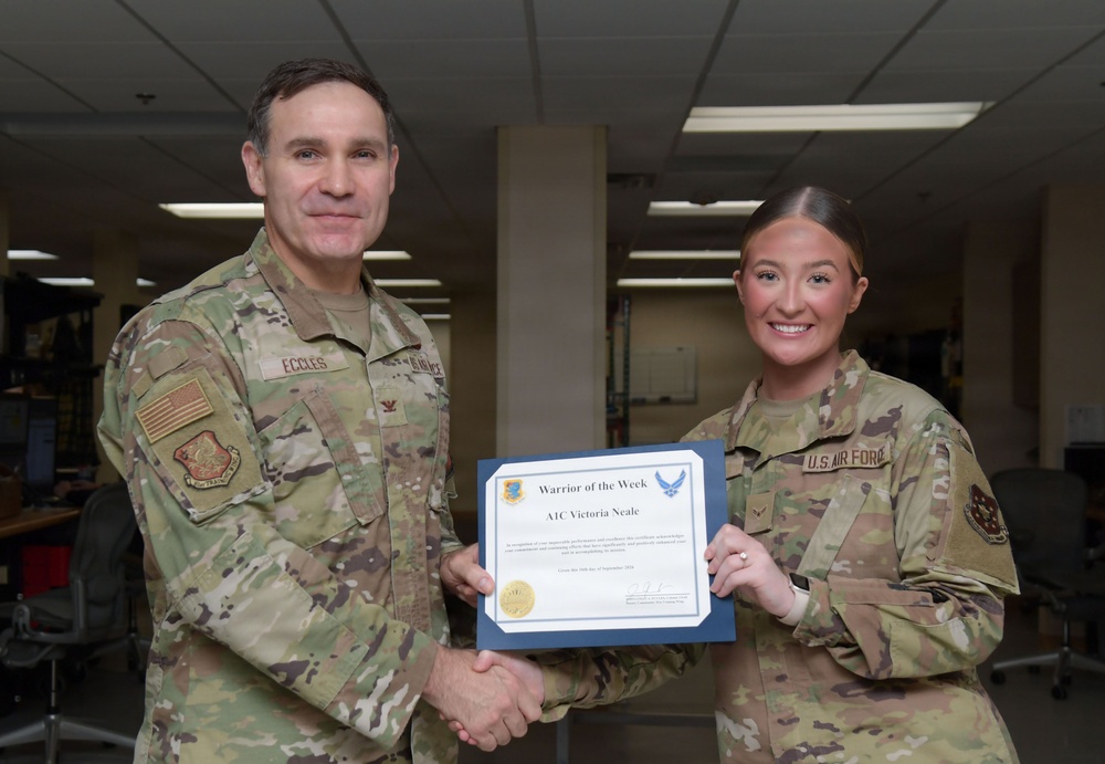Two U.S. Air Force personnel are posing together while holding a 'Warrior of the Week' certificate awarded to A1C Victoria Neale. The senior officer, identified by the name tape 'Eccles' and colonel rank insignia, is presenting the award with a handshake. The ceremony appears to take place in an office or administrative workspace.