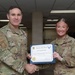 Two U.S. Air Force personnel are posing together while holding a 'Warrior of the Week' certificate awarded to A1C Victoria Neale. The senior officer, identified by the name tape 'Eccles' and colonel rank insignia, is presenting the award with a handshake. The ceremony appears to take place in an office or administrative workspace.