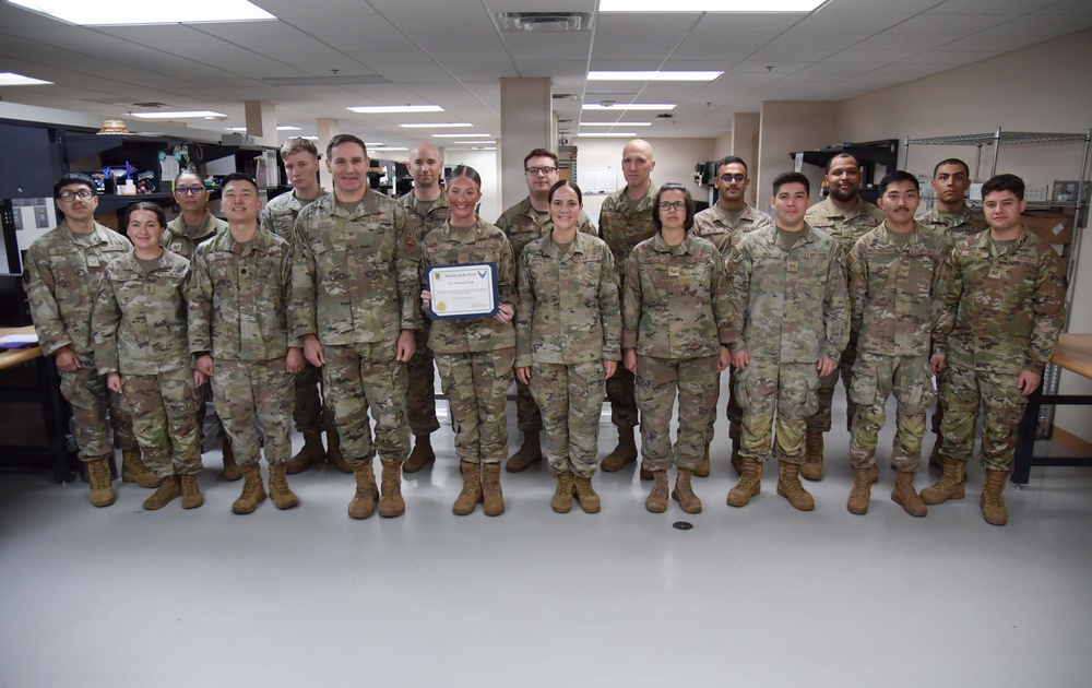 A group of approximately 18-20 military personnel in U.S. Army/Air Force Operational Camouflage Pattern (OCP) uniforms are posed together indoors for a formal group photo. One individual in the center is holding a certificate, suggesting a recognition or award ceremony, likely a 'Soldier/Airman of the Month/Year' or similar commendation. The setting appears to be an office or administrative workspace within a military installation.