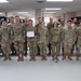 A group of approximately 18-20 military personnel in U.S. Army/Air Force Operational Camouflage Pattern (OCP) uniforms are posed together indoors for a formal group photo. One individual in the center is holding a certificate, suggesting a recognition or award ceremony, likely a 'Soldier/Airman of the Month/Year' or similar commendation. The setting appears to be an office or administrative workspace within a military installation.