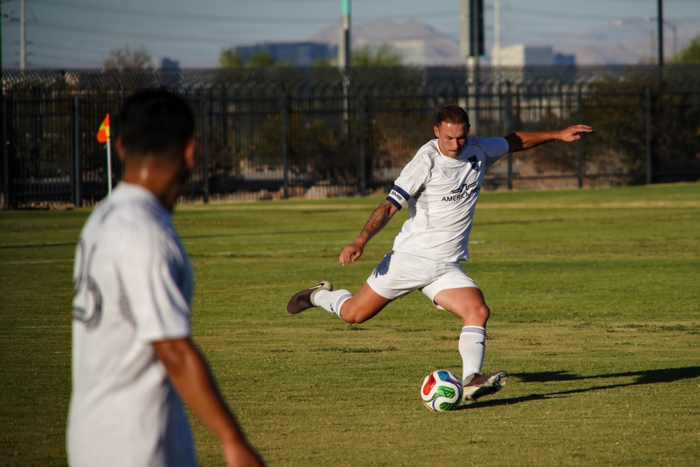 A soccer player in a white uniform is captured mid-kick, about to strike a soccer ball on a grass field. A teammate in a matching white uniform is visible in the foreground, blurred due to shallow depth of field. The setting appears to be an outdoor recreational or semi-professional soccer facility with fencing, a corner flag, and urban/mountain scenery in the background.