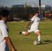 A soccer player in a white uniform is captured mid-kick, about to strike a soccer ball on a grass field. A teammate in a matching white uniform is visible in the foreground, blurred due to shallow depth of field. The setting appears to be an outdoor recreational or semi-professional soccer facility with fencing, a corner flag, and urban/mountain scenery in the background.