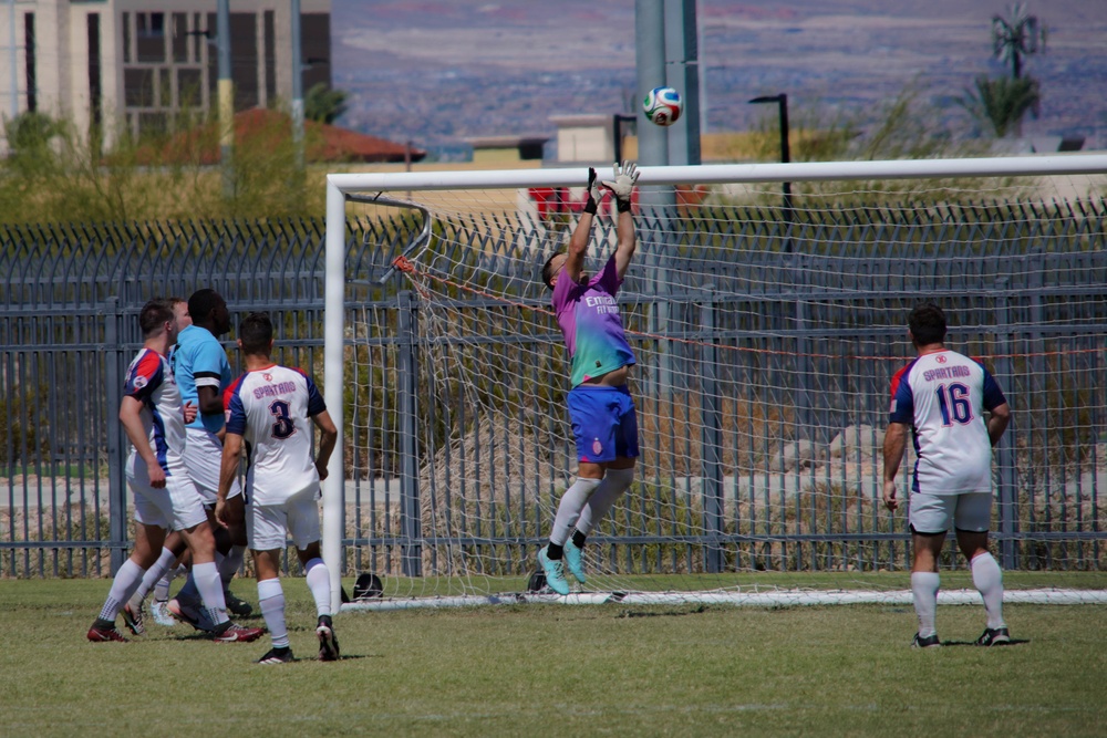 A soccer match is in progress with a goalkeeper in a purple and teal jersey leaping to make a save near the top of the goal. Several outfield players wearing white 'Spartans' jerseys with red and blue accents are positioned around the goal area. The match is being played on a grass field with a metal fence and desert landscape visible in the background.