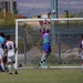 A soccer match is in progress with a goalkeeper in a purple and teal jersey leaping to make a save near the top of the goal. Several outfield players wearing white 'Spartans' jerseys with red and blue accents are positioned around the goal area. The match is being played on a grass field with a metal fence and desert landscape visible in the background.