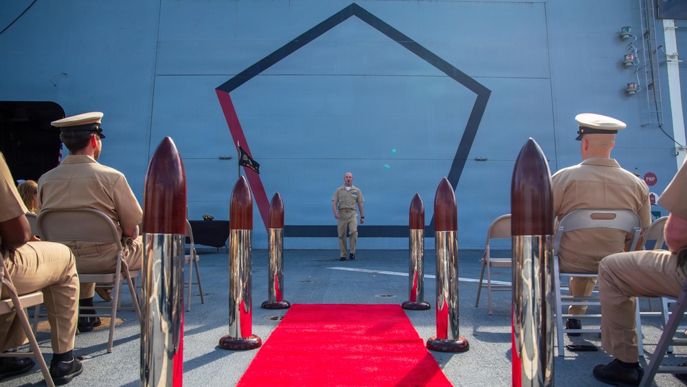A formal military ceremony is taking place on what appears to be the deck of a naval vessel. A single uniformed sailor walks down a red carpet flanked by decorative chrome-and-mahogany bollards, while seated naval personnel in khaki uniforms observe from both sides. A large pentagon-shaped emblem is painted on the ship's superstructure in the background.