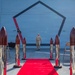 A formal military ceremony is taking place on what appears to be the deck of a naval vessel. A single uniformed sailor walks down a red carpet flanked by decorative chrome-and-mahogany bollards, while seated naval personnel in khaki uniforms observe from both sides. A large pentagon-shaped emblem is painted on the ship's superstructure in the background.