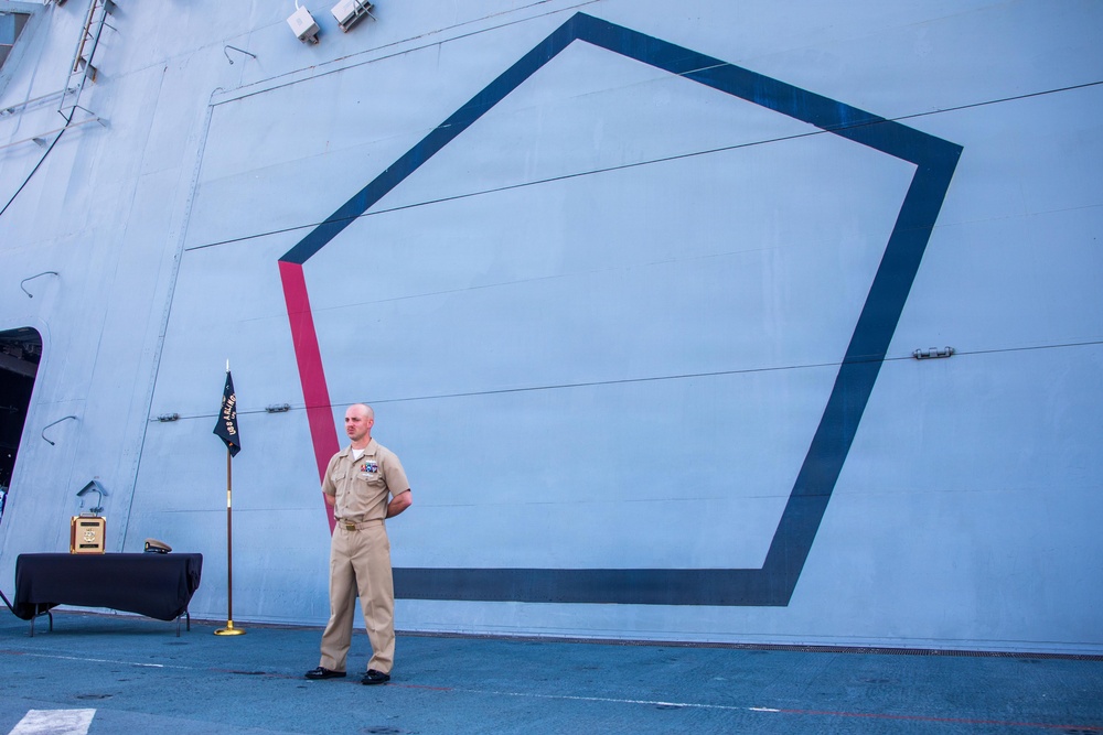 A U.S. Navy sailor in khaki service uniform stands at parade rest on the deck of a naval vessel, positioned in front of a large pentagon-shaped geometric design painted on the ship's superstructure. A unit or ship's commissioning pennant/flag on a gold stand is visible to his left, along with a draped table. The setting appears to be an official ceremony or formal event aboard the ship.