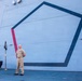 A U.S. Navy sailor in khaki service uniform stands at parade rest on the deck of a naval vessel, positioned in front of a large pentagon-shaped geometric design painted on the ship's superstructure. A unit or ship's commissioning pennant/flag on a gold stand is visible to his left, along with a draped table. The setting appears to be an official ceremony or formal event aboard the ship.