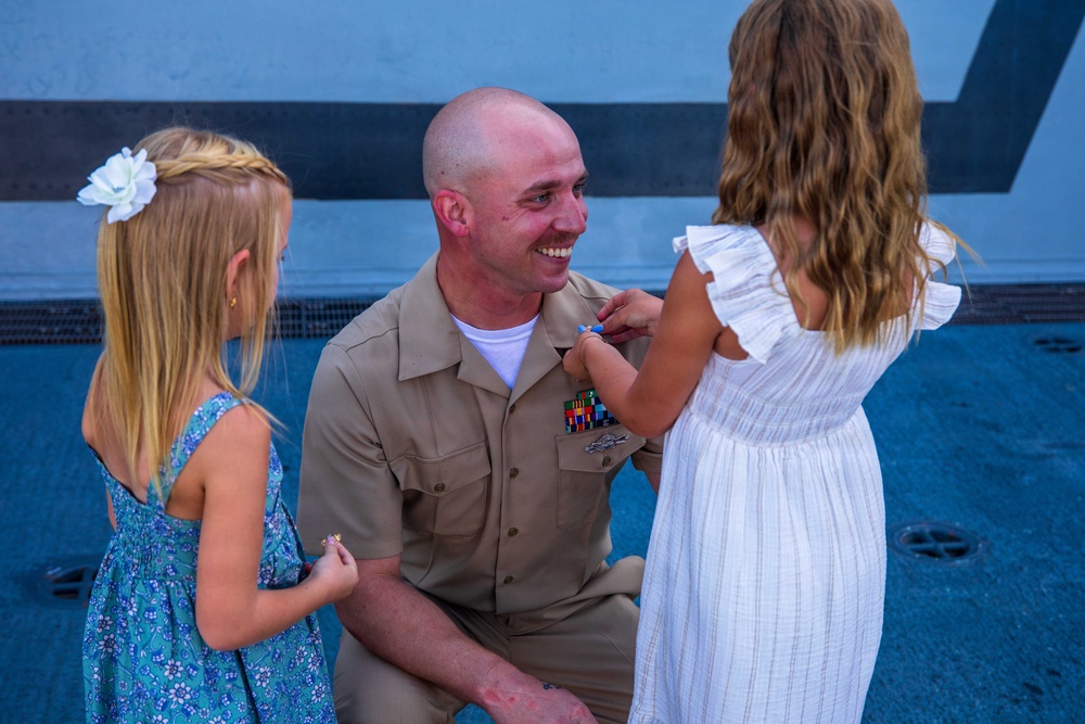 A smiling military serviceman in a tan/khaki uniform is kneeling down to the level of two young girls. One of the girls appears to be pinning or attaching something to his uniform near his ribbon rack. The scene appears to be a military promotion or award ceremony involving family members.
