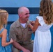A smiling military serviceman in a tan/khaki uniform is kneeling down to the level of two young girls. One of the girls appears to be pinning or attaching something to his uniform near his ribbon rack. The scene appears to be a military promotion or award ceremony involving family members.