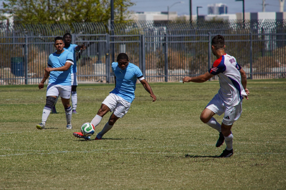 A soccer match is being played on a grass field, with players from two opposing teams in action. A player in a light blue uniform is in the process of kicking the ball while being challenged by a player in a white, red, and blue uniform. Two additional blue-uniformed players are visible in the background, one appearing to gesture or call out.