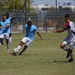 A soccer match is being played on a grass field, with players from two opposing teams in action. A player in a light blue uniform is in the process of kicking the ball while being challenged by a player in a white, red, and blue uniform. Two additional blue-uniformed players are visible in the background, one appearing to gesture or call out.