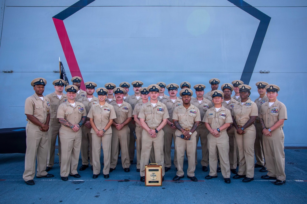 A formal group portrait of approximately 22-24 U.S. Navy personnel in khaki service uniforms (Service Uniform Charlie/Delta) posing in two rows. They are standing in front of what appears to be a large naval vessel or military structure featuring a distinctive geometric design with red and blue angular markings. A USN award plaque or trophy is displayed on the ground in front of the group.