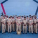 A formal group portrait of approximately 22-24 U.S. Navy personnel in khaki service uniforms (Service Uniform Charlie/Delta) posing in two rows. They are standing in front of what appears to be a large naval vessel or military structure featuring a distinctive geometric design with red and blue angular markings. A USN award plaque or trophy is displayed on the ground in front of the group.