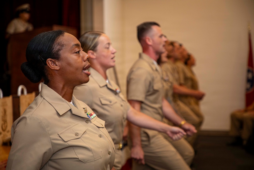 A group of U.S. Navy personnel in tan/khaki service uniforms are seated in a row, appearing to be singing or reciting something in unison during a formal military ceremony or event. A figure in uniform is visible at a podium in the background, and a flag is partially visible on the right side. The foreground subject displays service ribbons on her uniform.