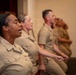 A group of U.S. Navy personnel in tan/khaki service uniforms are seated in a row, appearing to be singing or reciting something in unison during a formal military ceremony or event. A figure in uniform is visible at a podium in the background, and a flag is partially visible on the right side. The foreground subject displays service ribbons on her uniform.