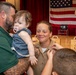 A U.S. Navy promotion or pinning ceremony is taking place in what appears to be a military hall. A female sailor in khaki uniform is being pinned by a child while her family — a man holding a baby girl with a pacifier — looks on warmly. An American flag and engraved wooden plaques are visible in the background.
